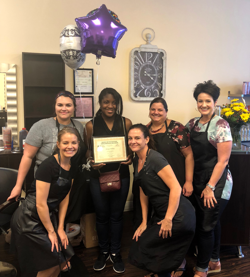 Young woman posed with a program completion certificate, surrounded by her hair salon coworkers.